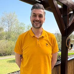 Mann lächelt im gelben Polohemd vor Holzbalkon mit Natur im Hintergrund.