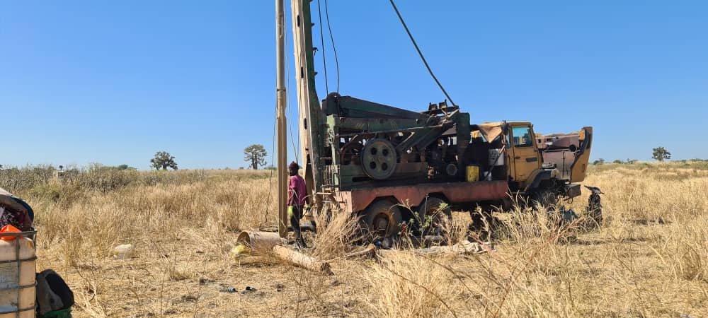 Eine Person steht neben einer alten Bohrmaschine auf trockenem gelben Feld unter blauem Himmel
