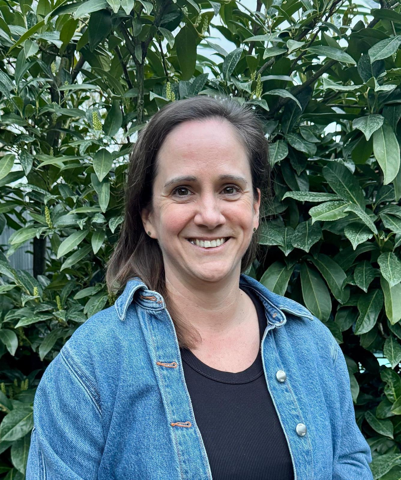 Eine braunhaarige Frau mit freundlichem Lächeln steht vor grünen Blättern