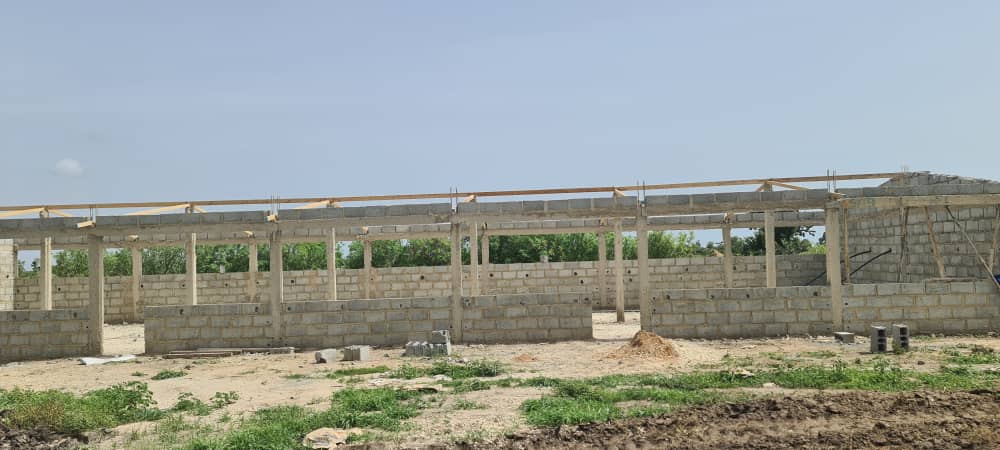 Ein rohbauähnliches Gebäude aus grauen Ziegeln steht auf sandigem Boden mit wenig Vegetation