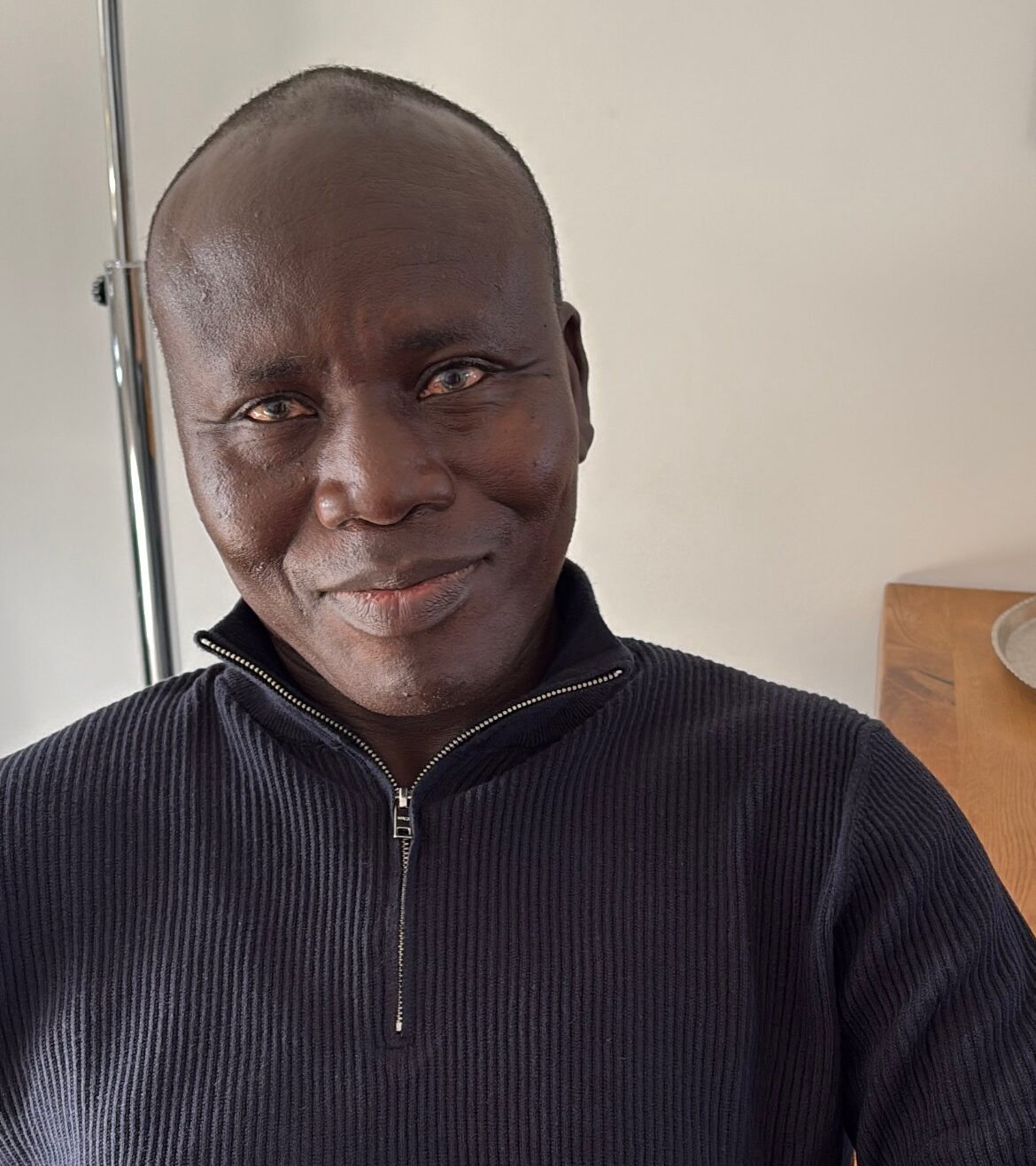 Ein lächelnder Mann mit dunkler Haut trägt einen schwarzen Pullover und sitzt entspannt