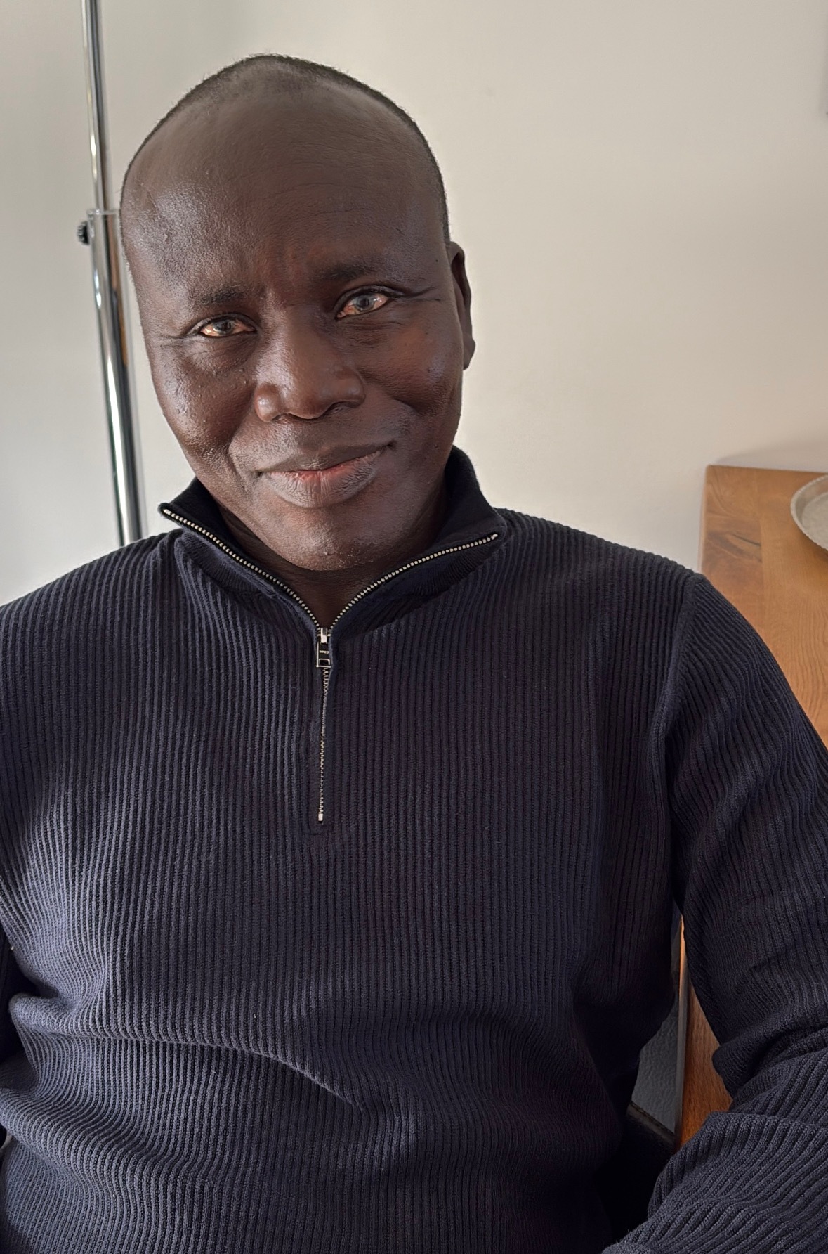 Ein lächelnder Mann mit dunkler Haut trägt einen schwarzen Pullover und sitzt entspannt